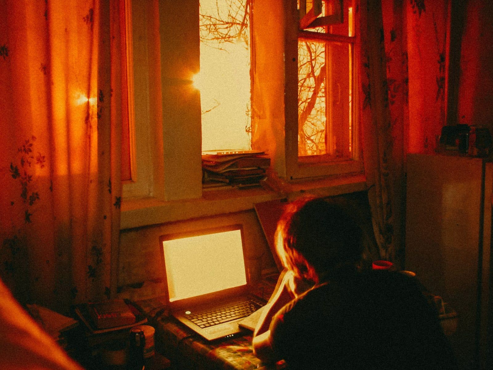 A person at a laptop in a warm, lamp-lit room at dawn